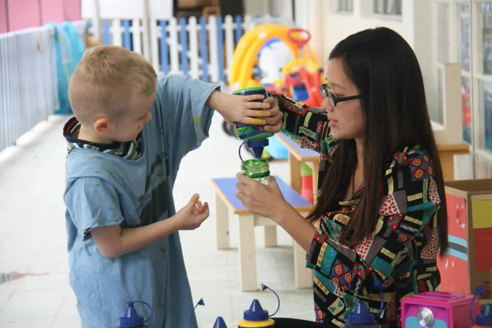 Teacher helping a preschool child pour paint into a container.
