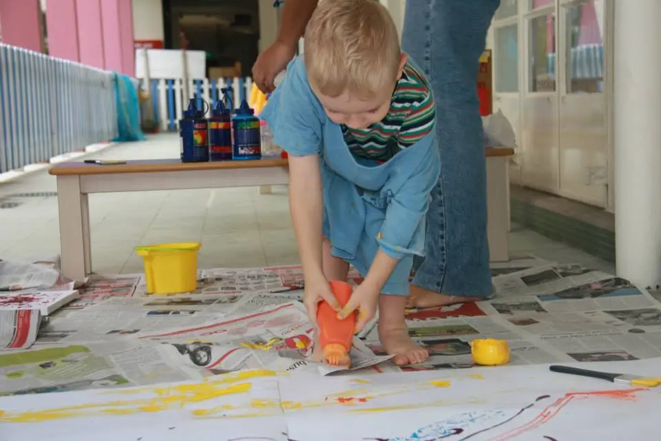 Young child squeezing orange paint onto a large sheet of paper.